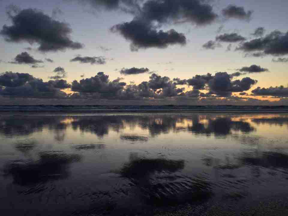Scattered dark clouds above cumulus clouds over the ocean, waves breaking upon a wet shore, reflecting the clouds and post-sunset sky, bits of orange in the gaps between the clouds on the horizon.