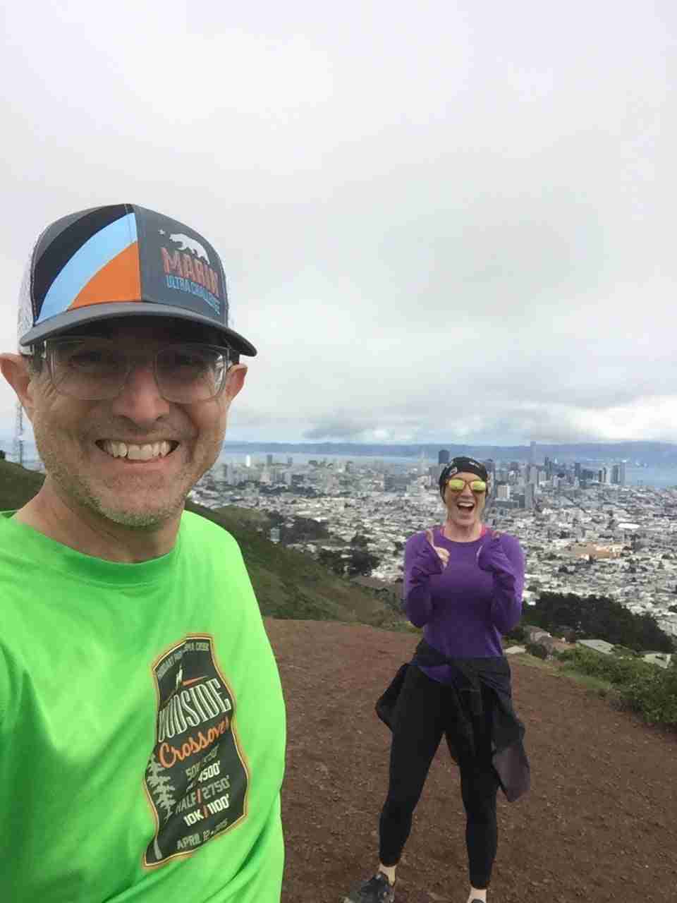 Tantek and Courtney smiling, more than six feet apart from each other, on top of the North Peak of Twin Peaks, with downtown San Francisco in the distance behind them.