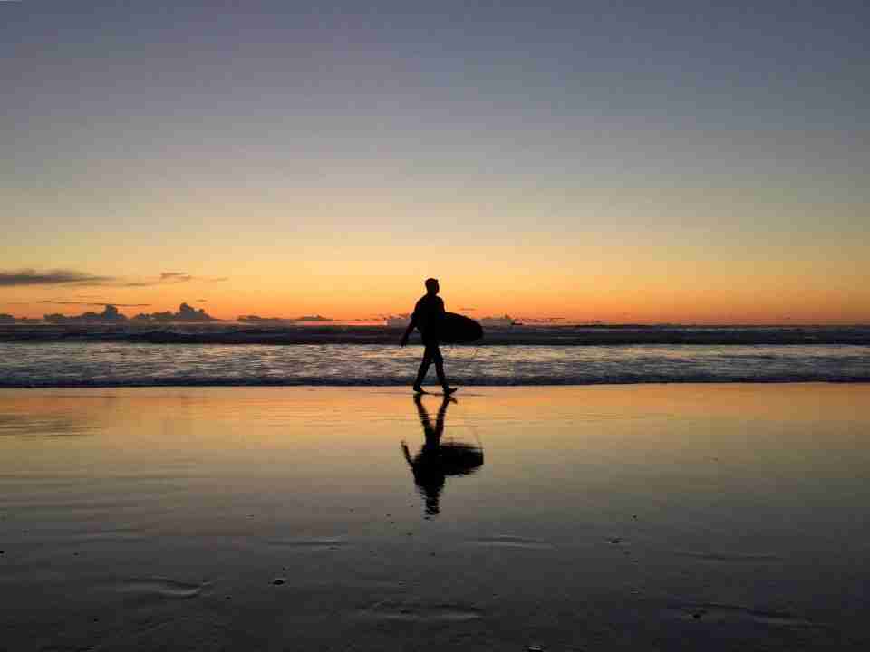 A surfer walking out of the surf with their surfboard tucked under their arm, just after sunset, clear skies, yellow orange horizon over the ocean, wet sand reflecting the sky and surfer.