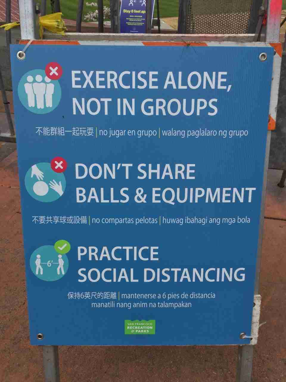 Close-up of a sign with three statements, each translated into Chinese, Spanish, and Filipino, stating “EXERCISE ALONE, NOT IN GROUPS”, “DON’T SHARE BALLS & EQUIPMENT”, and “PRACTICE SOCIAL DISTANCING”, below those a small San Francisco Recreation & Parks logo.