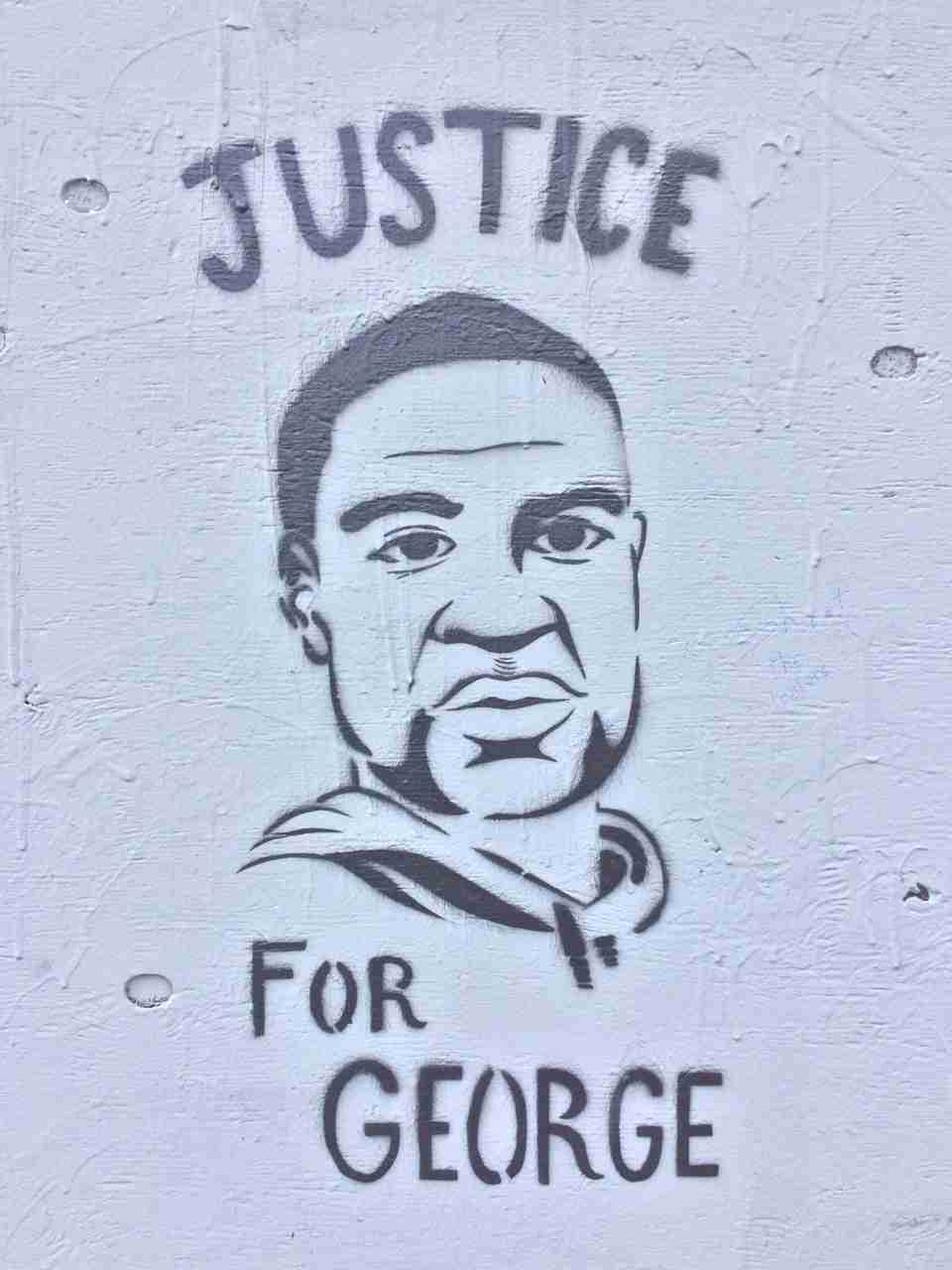 Stencil street art of George Floyd on a gray wall with the word JUSTICE above his head, and FOR GEORGE below. Two of the dried drippings of gray paint appear as if they are tears coming from his right eye.