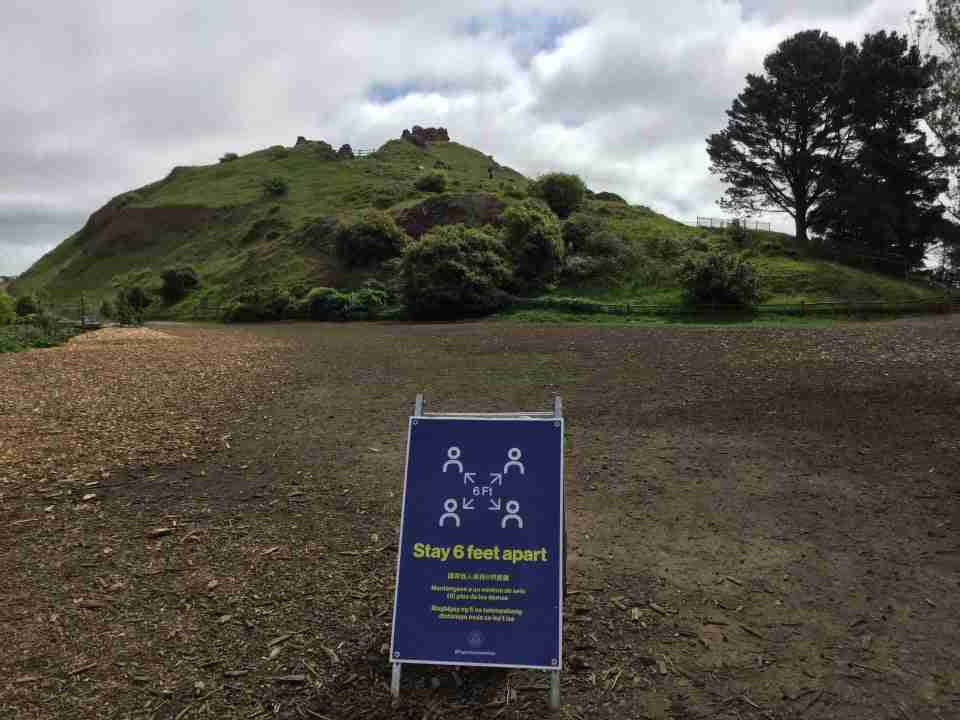 Stay 6 feet apart sign in the dirt field at the base of Corona Heights Park, the hill in the middle green from recent rains, bits of blue sky peaking through white clouds overhead.