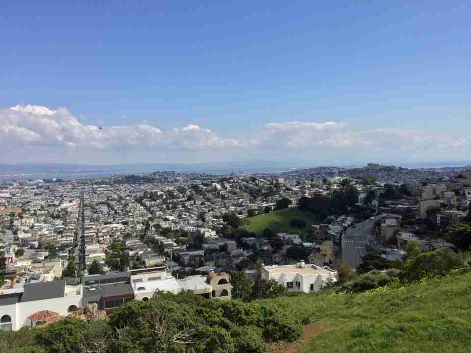 Blue skies with a small band of low hanging white puffy clouds in the distance over a clear view of East Bay mountains, the bay itself with tiny boats & ships, Bernal Heights Park, many San Francisco neighborhoods, and the green grass & a few bushes nearby from the side of Tank Hill Park.