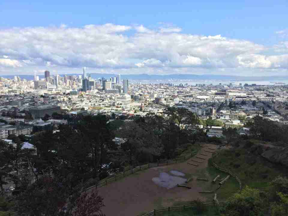 Light blue skies, low hanging scattered white gray clouds over East Bay hills in the distance, downtown San Francisco, and trees, green bushes, grass, & dirt paths of Corona Heights Park below, with a couple of large puddles from recent rains.