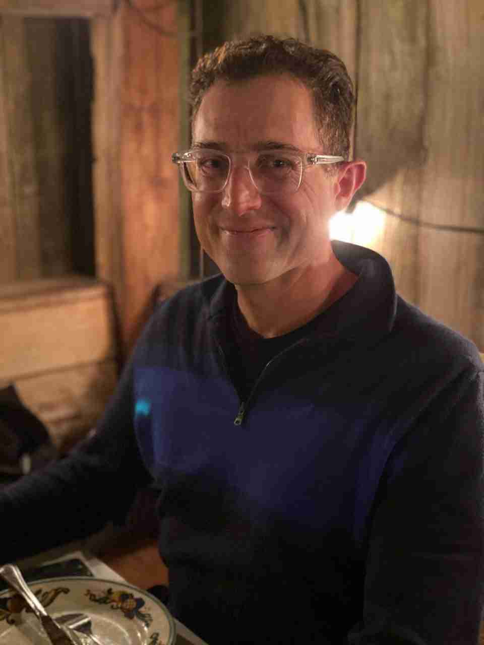 Tantek smiling in the Zazie patio in the evening, lit by lamps, wearing a half-zip dark blue sweater with a medium blue wide stripe across it.