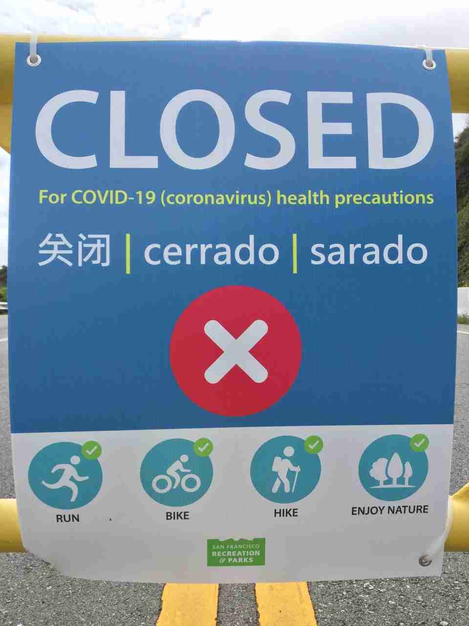 Close-up of a sign stating CLOSED For COVID-19 (coronavirus) health precautions, translations in Chinese & Spanish, a big red circle with a white x in the middle, and below that a row of four icons of activities with checkmarks (white on green background) in their top right corners, for run, bike, hike, and enjoy nature, below those a small San Francisco Recreation & Parks logo.